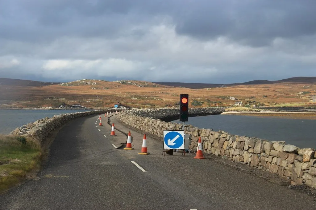 Roadworks with cones and a temporary traffic light on a stone causeway across a Highland loch, laying fibre-optic cable