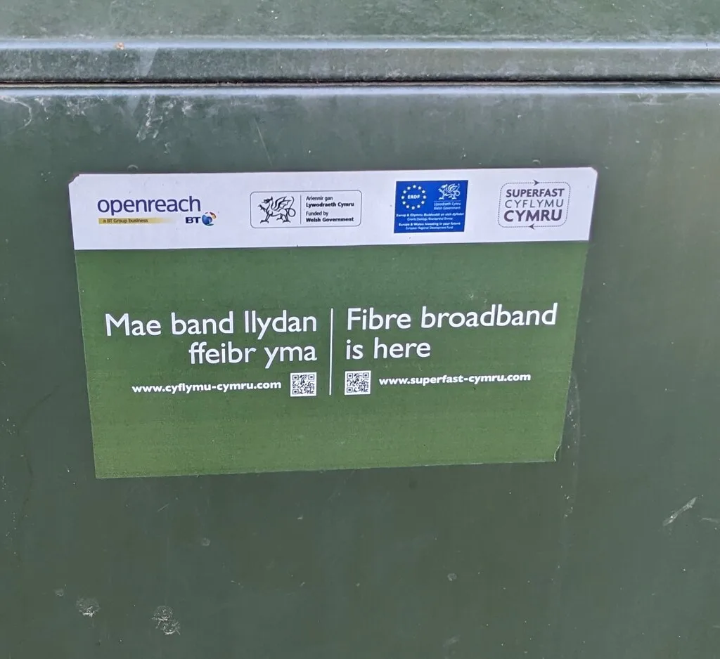 A green street cabinet with a bilingual Welsh and English sticker reading Fibre broadband is here